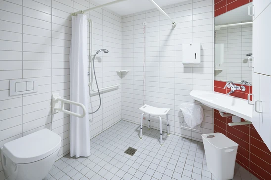 Salle de bain adapt&eacute;e PMR avec douche &agrave; l'italienne &eacute;quip&eacute;e de barres d'appui murales blanches, si&egrave;ge de douche rabattable et tabouret blanc r&eacute;glable
