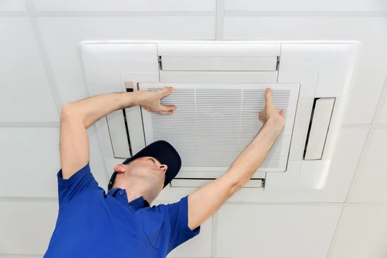 Un technicien en uniforme bleu et casquette noire installe ou remplace un filtre &agrave; air dans un syst&egrave;me de ventilation plafond int&eacute;rieur blanc