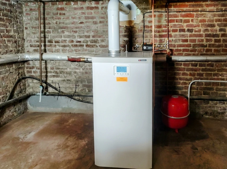 Un technicien en salopette et chemise à carreaux effectue la maintenance d'une chaudière murale domestique dans un local technique