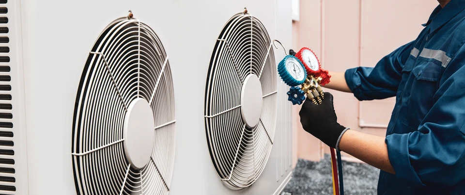 Technicien en climatisation travaillant dans une pièce résidentielle, portant une combinaison rouge et gants bleus, manipulant des outils près d'une unité de climatisation murale ouverte